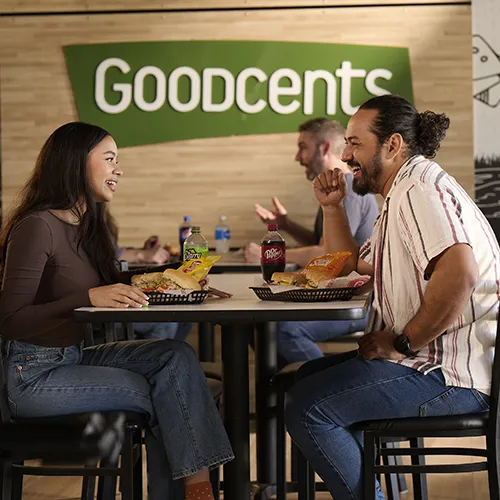 A couple of people enjoying a meal in front of a Goodcents sign
